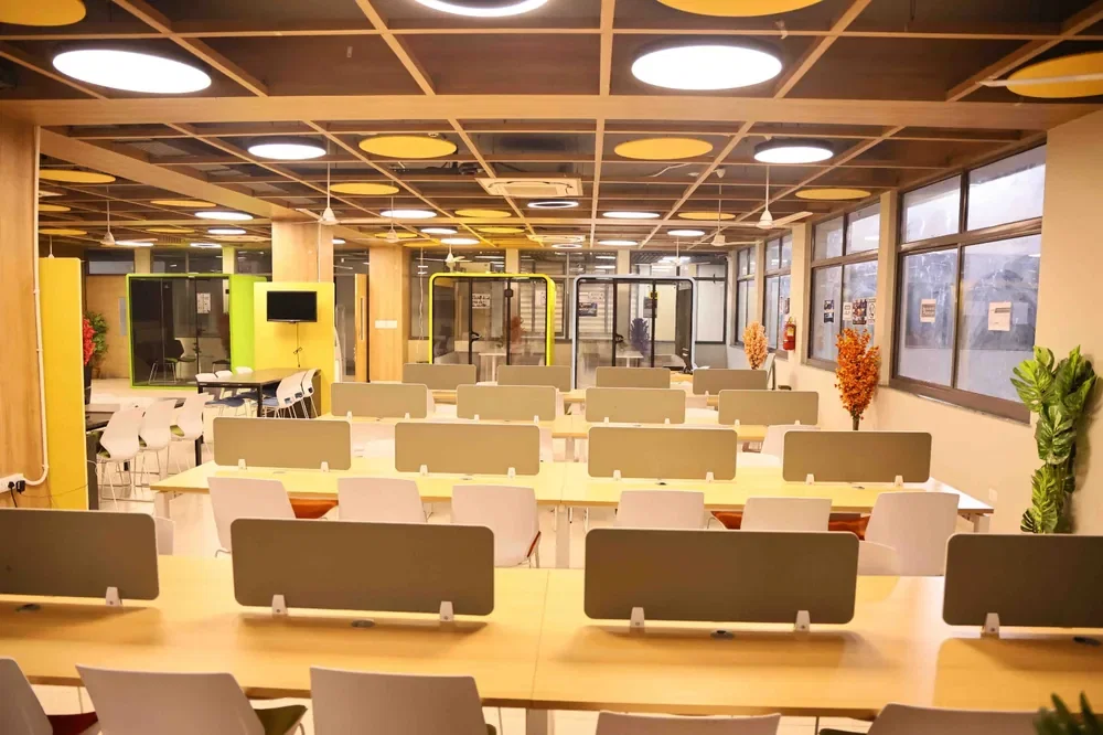 Rows of shared desks under a timber grid ceiling, phone booths along one side